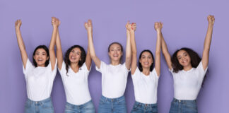 Für Emanzipation und Gleichberechtigung Foto zeigt lachende Frauen mit hochgestreckten Armen vor lilafarbenen Hintergrund