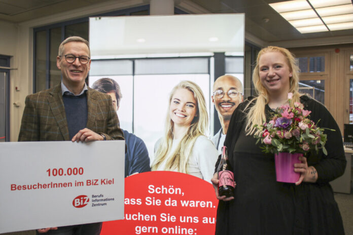 Hans-Martin Rump, Leiter der Agentur für Arbeit Kiel, überreicht der Kundin einen Blumenstrauß und eine Flasche alkoholfreien Rosmarin-Prosecco.