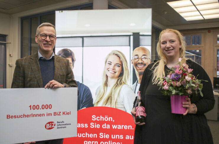 Hans-Martin Rump, Leiter der Agentur für Arbeit Kiel, überreicht der Kundin einen Blumenstrauß und eine Flasche alkoholfreien Rosmarin-Prosecco.