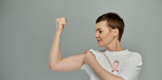 Sport bei Krebs: KITT sucht Teilnehmende für Teletherapie-Studie A woman with short hair flexes her bicep, displaying a pink ribbon symbolizing breast cancer awareness.
